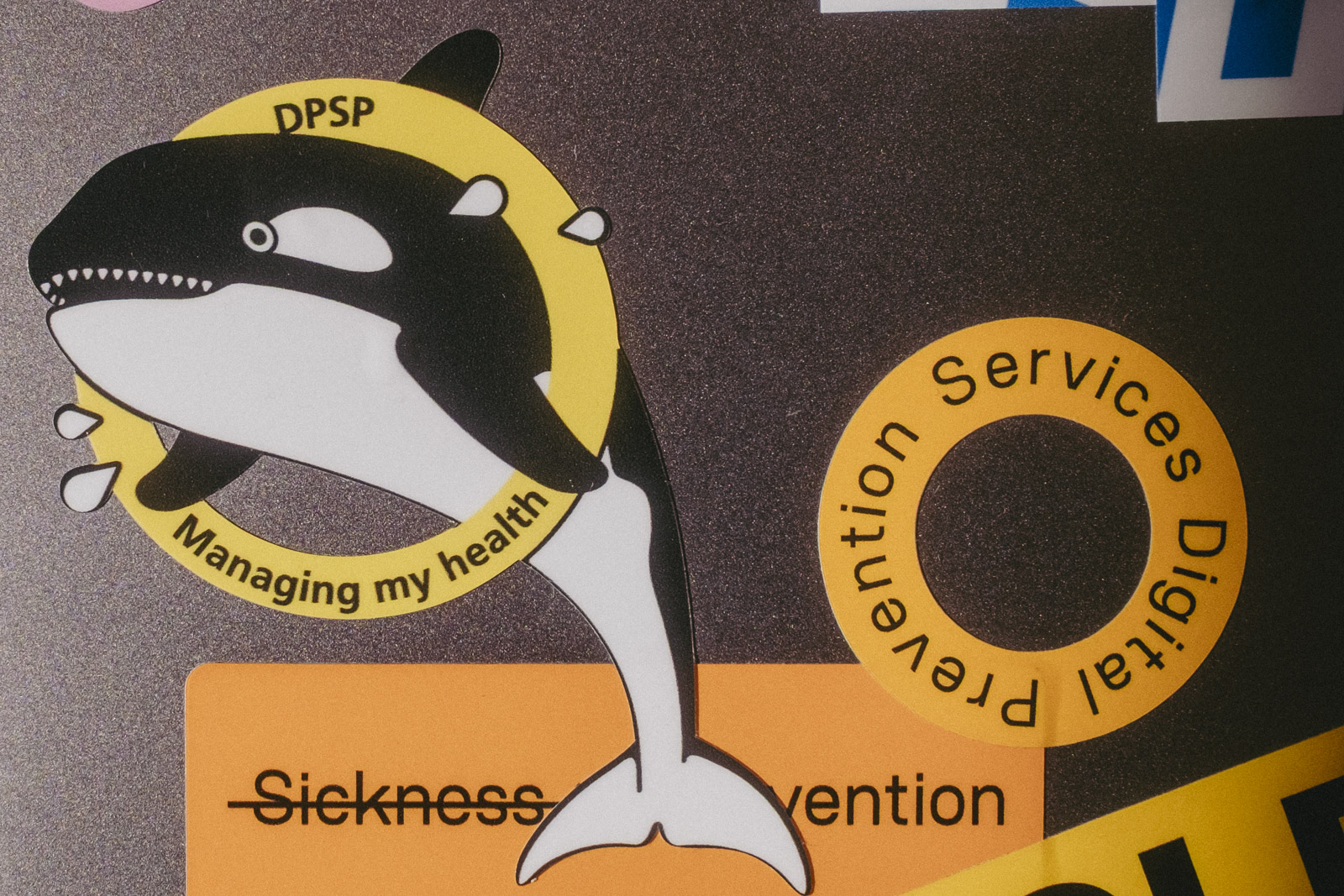 Stickers on a laptop focusing a mission patch showing a sweaty orca jumping through a circle with the words 'DPSP' and 'Managing my health'